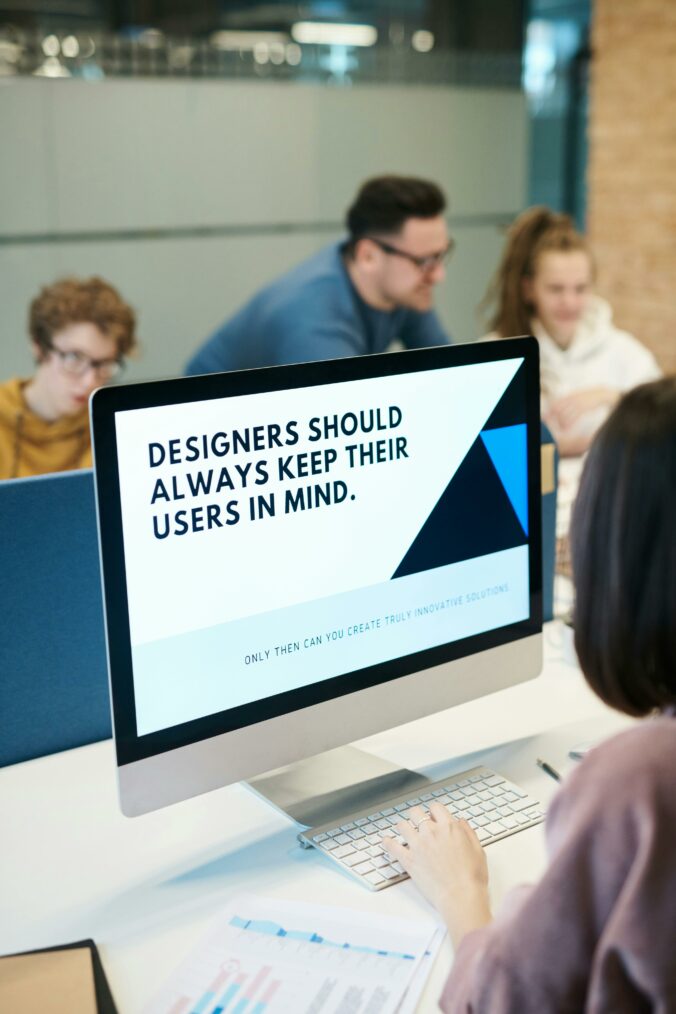 A computer screen reads "Designers should always keep their users in mind." In the background, a teacher helps a student while another student works independently.