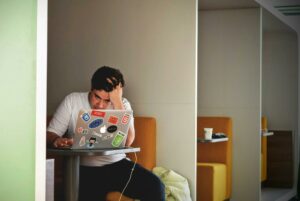 A young man sits in front of a laptop with his head in his hand.